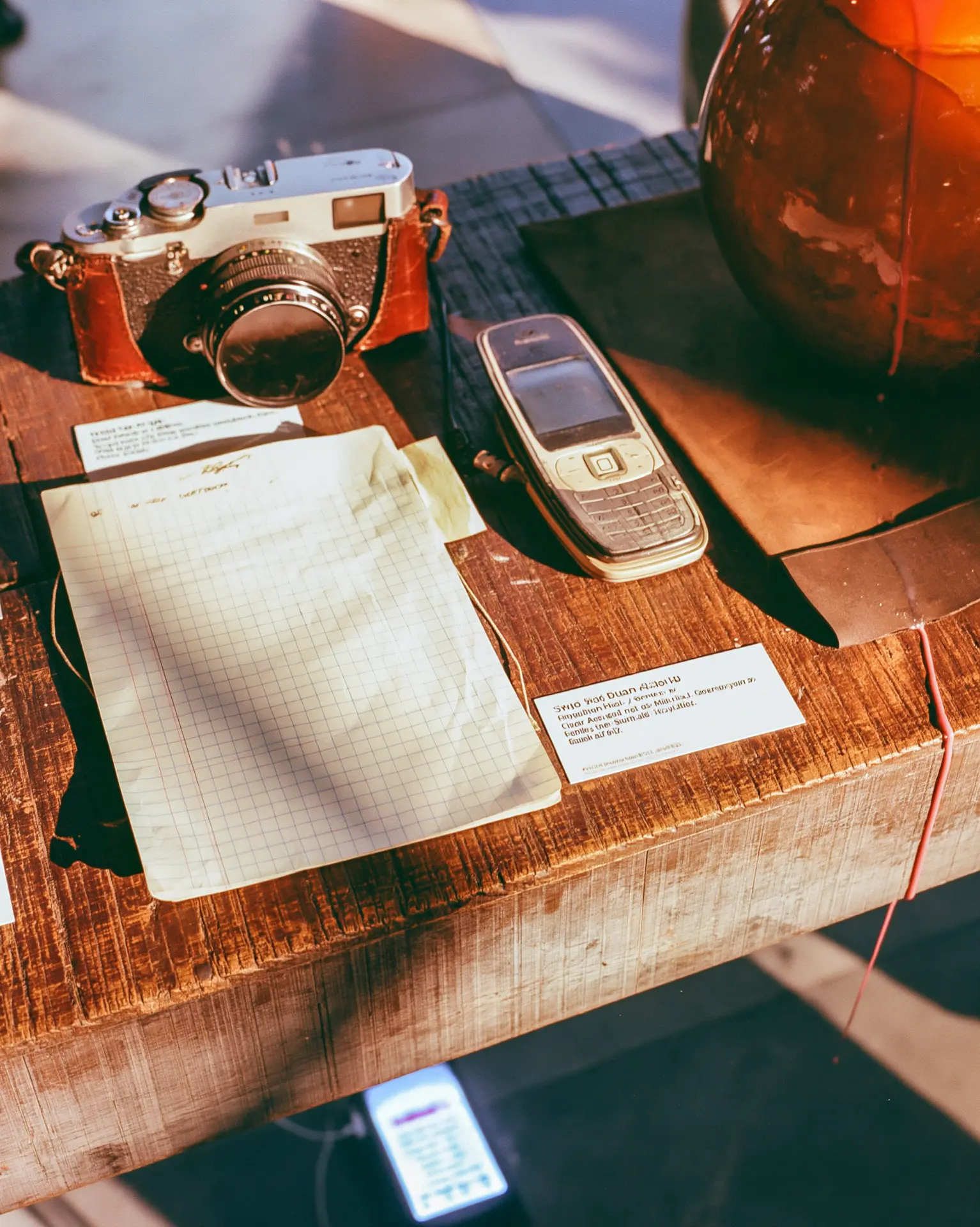 a vintage film camera, a scuffed flip phone and a grid-paper notebook arranged like museum artifacts on a wooden plinth with printed placards, warm afternoon light
