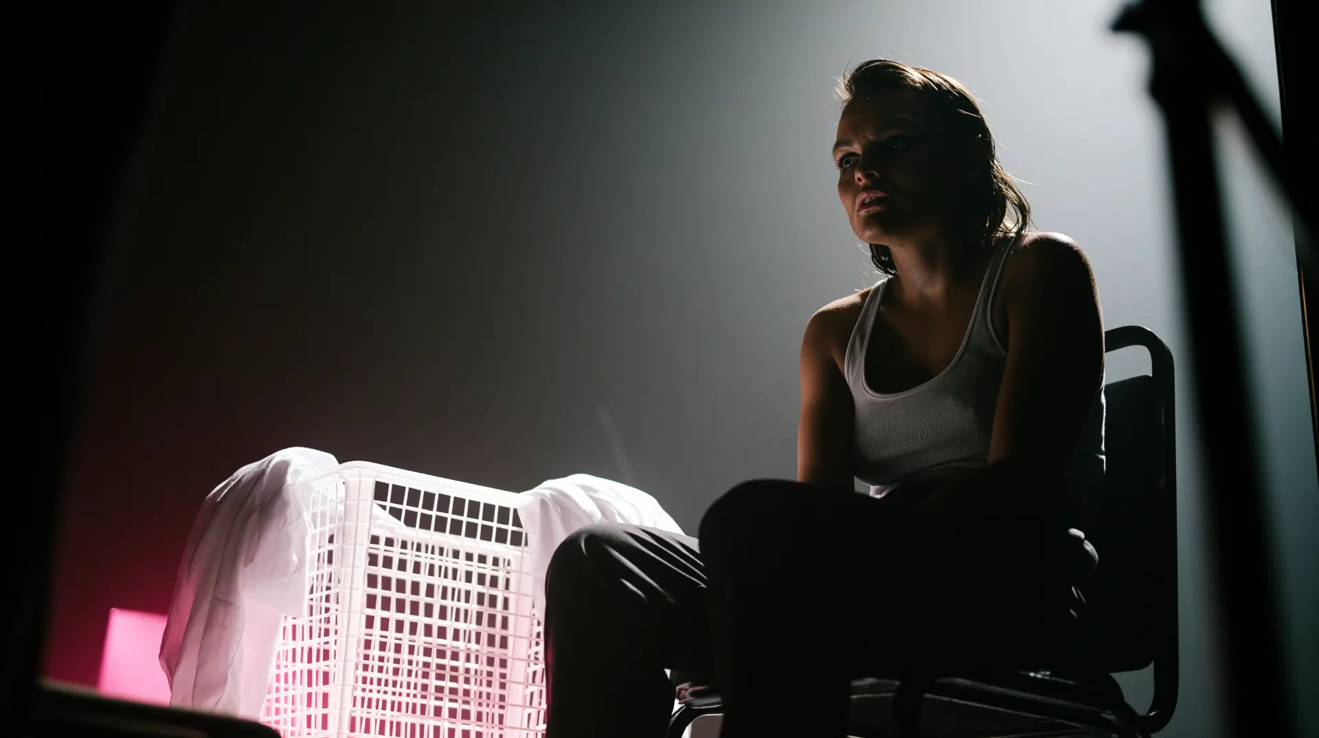 a lone figure in a white tank top sitting on a plain chair in a dark studio, backlit like a documentary confessional interview, a laundry basket beside her glowing pink